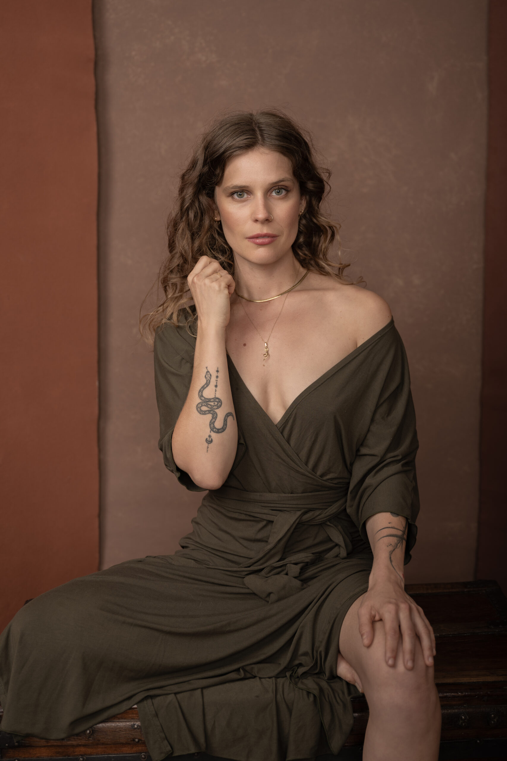 Portrait of a woman with wavy hair wearing and off-shoulder olive dress, showcasing a snake tattoo on her arm, against a warm earthy background.